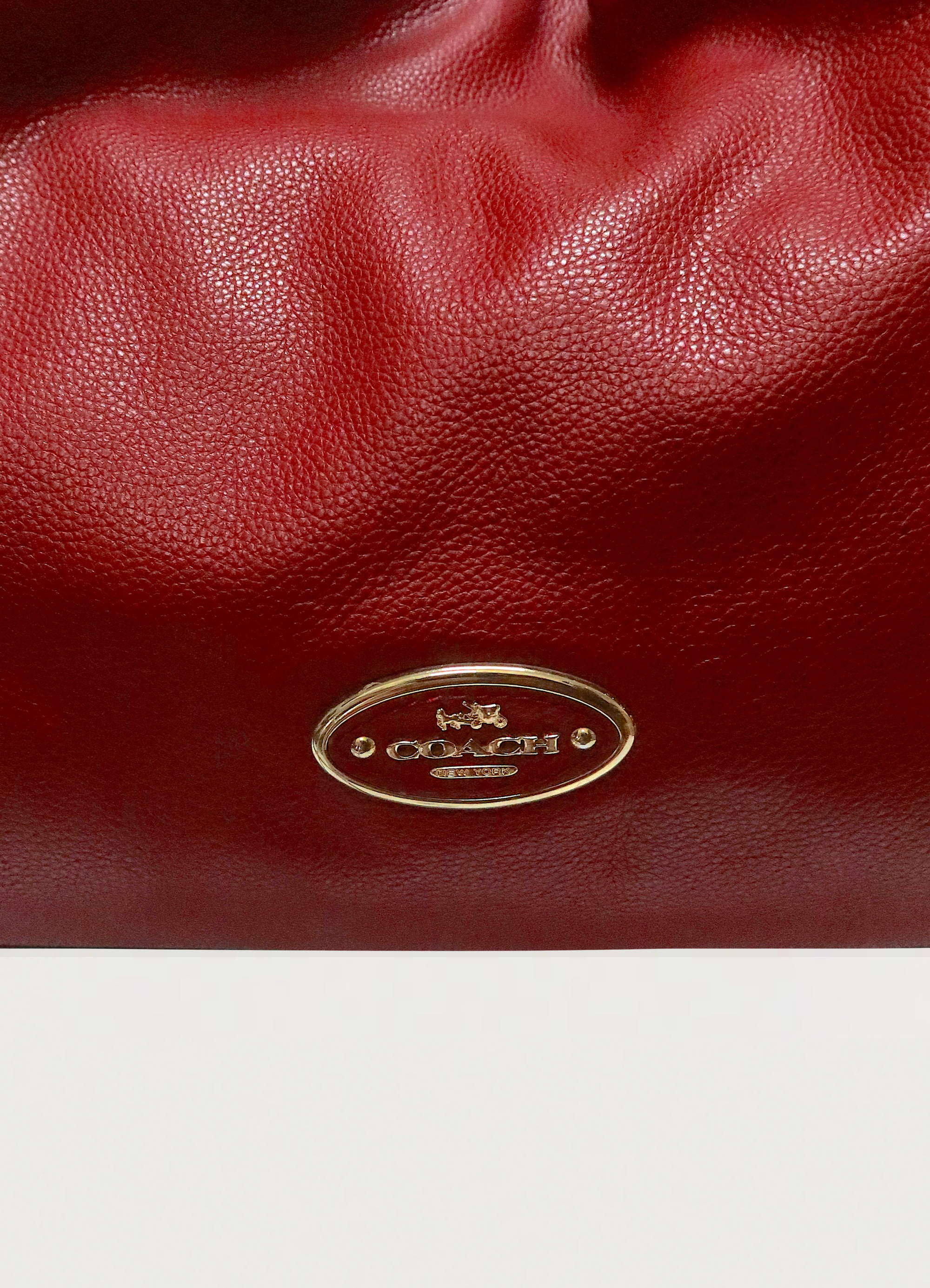 Close up of the gold-tone Coach oval logo plaque on red pebble leather